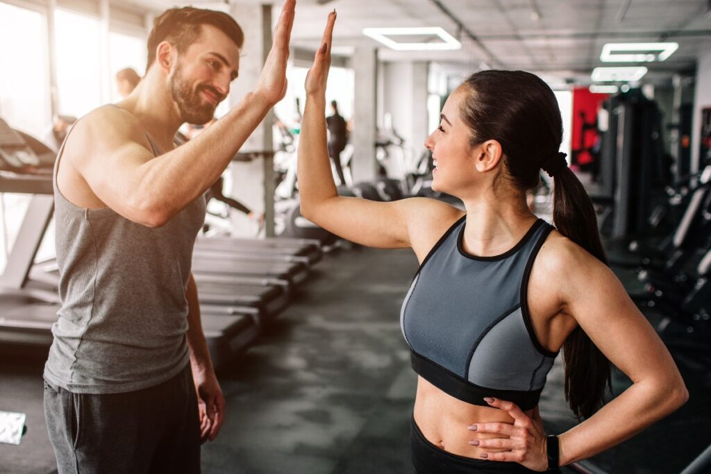 A beautiful girl and her well-built boyfriend are greeting each other with a high-five. They are happy to see each othr in the gym. Young people are ready to start their workout.